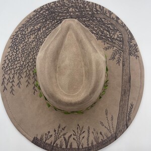 May include: A tan fedora-style hat with a wide brim. The brim features a hand-drawn tree and foliage design in dark brown. A green leafy band encircles the base of the crown. The hat is a stylish accessory.