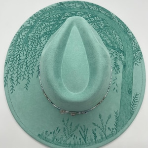 May include: A mint green fedora-style hat with a forest scene design. The hat features a detailed illustration of trees, foliage, and a patterned band. The hat's brim is adorned with a nature-inspired design, creating a unique accessory.