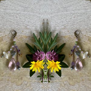 May include: A pair of dangle earrings featuring faceted pink teardrop crystals, small silver-toned heart charms, and an assortment of purple, green, and clear beads on thin wires. The earrings are arranged symmetrically with fresh flowers.