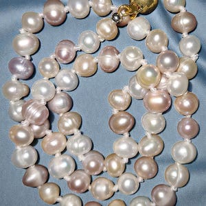 May include: A multi-strand pearl necklace featuring a variety of colors, including white, pink, and peach. The pearls are irregularly shaped and strung on a white cord, with a gold-colored clasp.