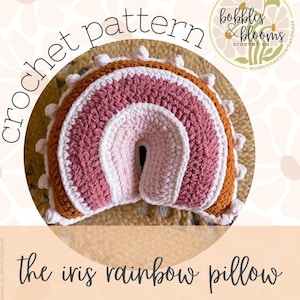 May include: A crocheted rainbow pillow in shades of pink, white, and brown. The pillow has a rounded shape with a central opening. The text "crochet pattern" and "the iris rainbow pillow" are displayed. The logo for "bobbles & blooms crochet co." is also visible.