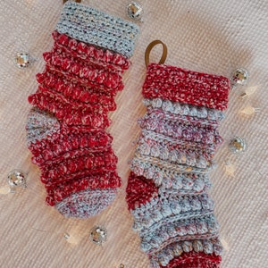 May include: Two crocheted Christmas stockings in a red and blue colour scheme. The stockings have a textured, puffy design and are decorated with small white pom-poms.