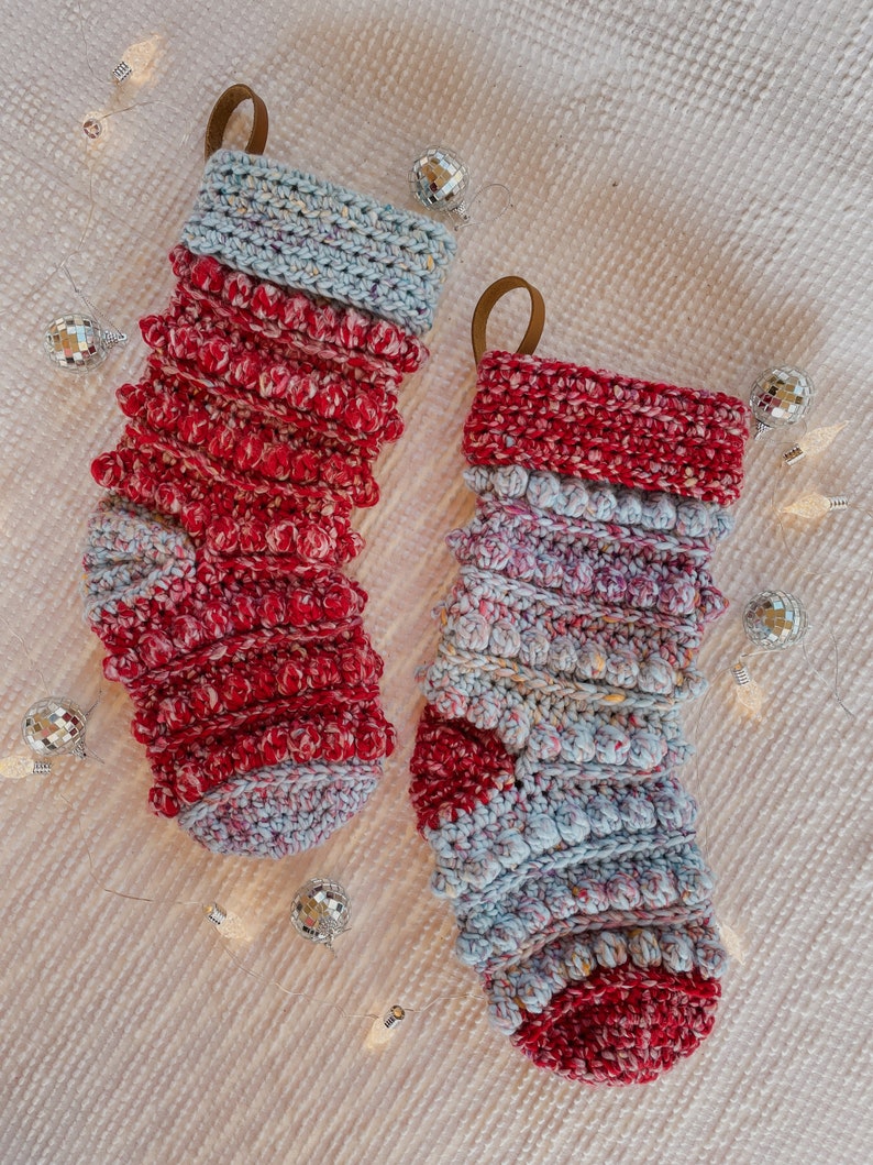 May include: Two crocheted Christmas stockings in a red and blue colour scheme. The stockings have a textured, puffy design and are decorated with small white pom-poms.