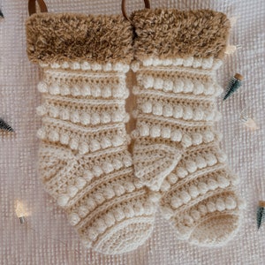 May include: Two white crocheted Christmas stockings with brown faux fur trim and a brown leather loop at the top.