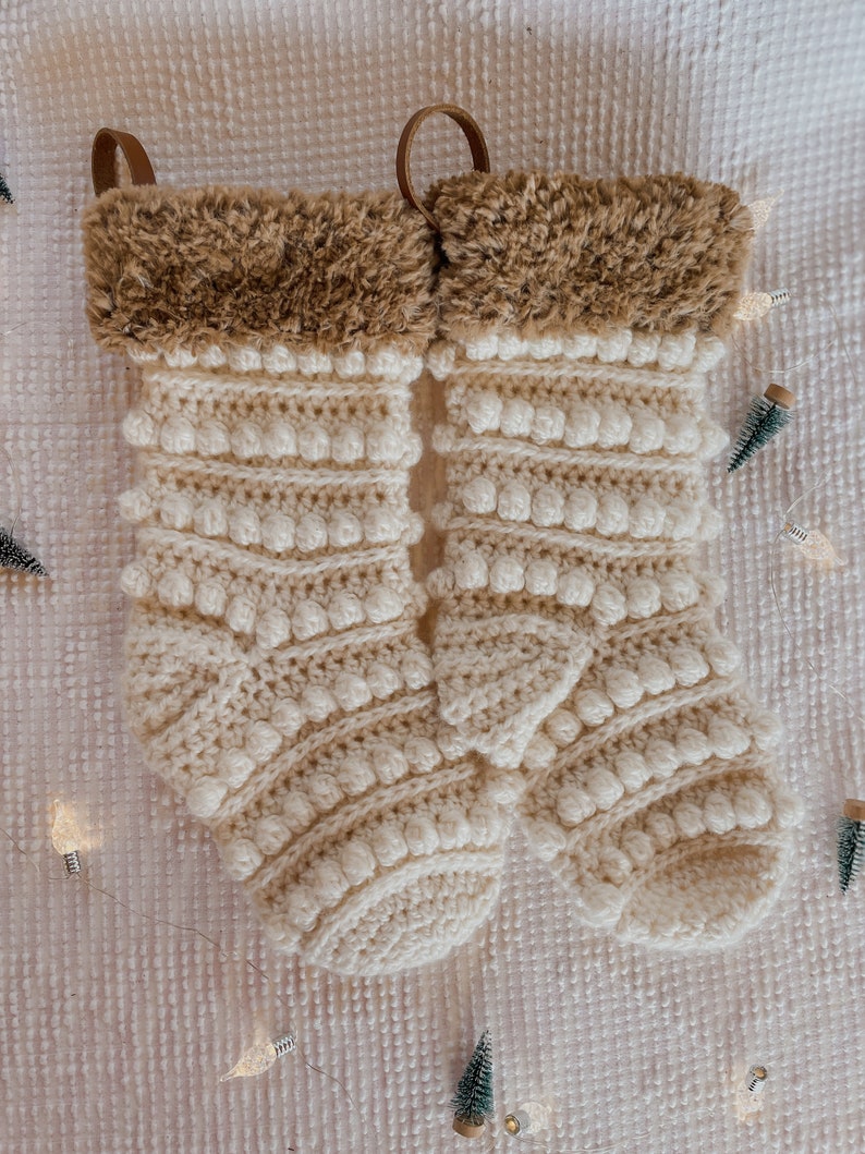 May include: Two white crocheted Christmas stockings with brown faux fur trim and a brown leather loop at the top.