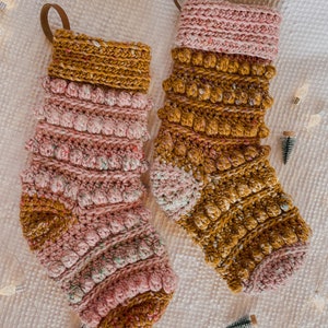 May include: Two crocheted Christmas stockings in a pink and mustard yellow colour scheme. The stockings feature a textured, bobble stitch design.