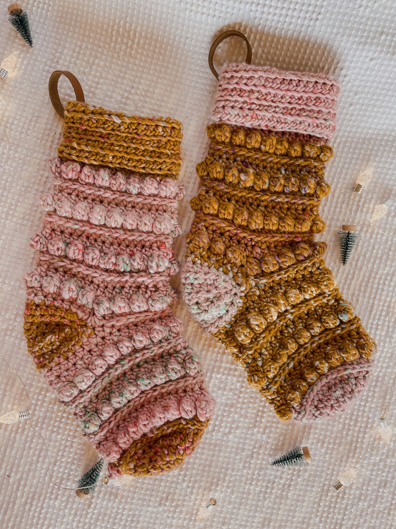 May include: Two crocheted Christmas stockings in a pink and mustard yellow colour scheme. The stockings feature a textured, bobble stitch design.