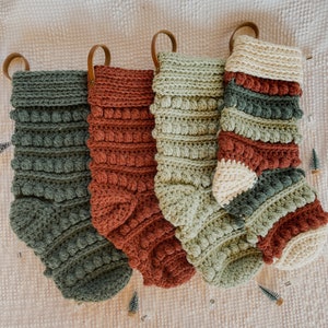 May include: Four crocheted Christmas stockings in green, red and white with brown leather loops. The stockings are decorated with a textured, bobble stitch.