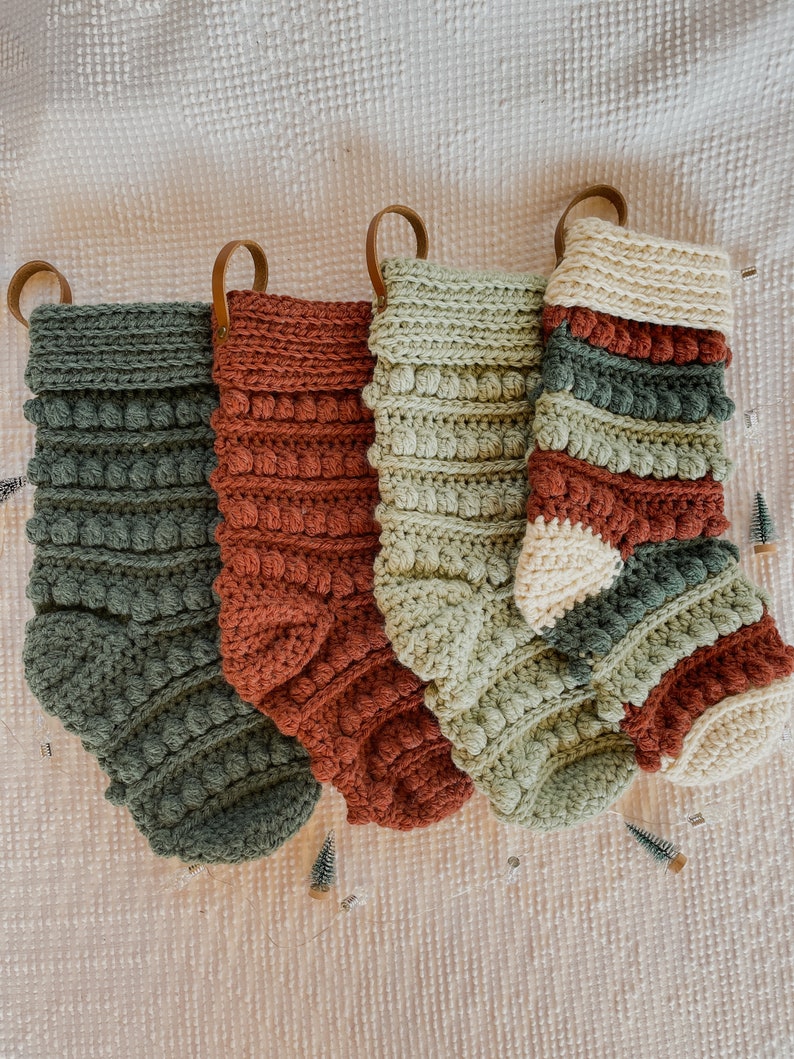 May include: Four crocheted Christmas stockings in green, red and white with brown leather loops. The stockings are decorated with a textured, bobble stitch.