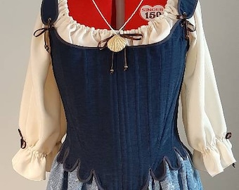 May include: A blue and white floral print dress with a blue bodice and a white blouse with puffed sleeves. The bodice has a scalloped hem and is cinched at the waist. The dress is accessorized with a blue bow and a seashell necklace.