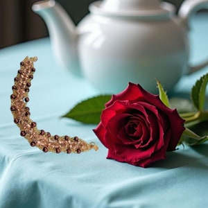 May include: A crescent-shaped beaded bracelet featuring small, light-colored crystals and dark, round beads arranged in a curved pattern, positioned next to a single, vibrant red rose with green leaves and a white ceramic teapot in the background.