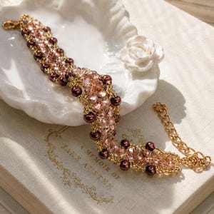 May include: A beaded bracelet featuring a woven pattern of light pink faceted crystals, dark brown round pearls, and small gold-toned seed beads. The bracelet includes a gold-toned metal chain extender with a lobster claw clasp at one end.