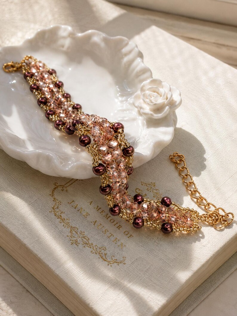 May include: A beaded bracelet featuring a woven pattern of light pink faceted crystals, dark brown round pearls, and small gold-toned seed beads. The bracelet includes a gold-toned metal chain extender with a lobster claw clasp at one end.