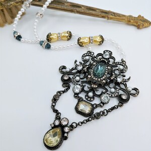 May include: An ornate, antique-style necklace with a large central pendant featuring a teal-colored oval stone. The pendant is surrounded by clear crystals and small faux pearls. The necklace has a chain and a pearl strand.