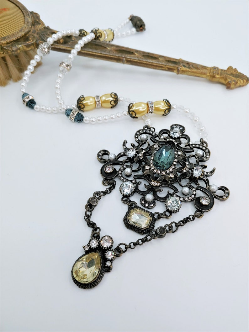 May include: An ornate, antique-style necklace with a large central pendant featuring a teal-colored oval stone. The pendant is surrounded by clear crystals and small faux pearls. The necklace has a chain and a pearl strand.
