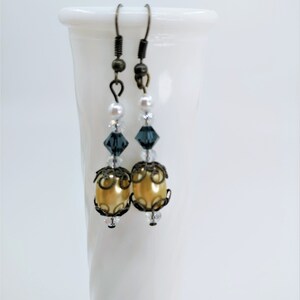 May include: A pair of dangle earrings with a vintage aesthetic. Each earring features a gold-colored, oval-shaped bead, accented with dark metal filigree and clear crystals. Above the gold bead are dark blue crystals, small white pearls, and a dark metal hook.