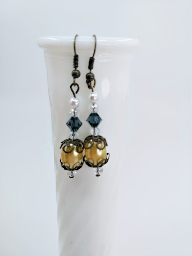 May include: A pair of dangle earrings with a vintage aesthetic. Each earring features a gold-colored, oval-shaped bead, accented with dark metal filigree and clear crystals. Above the gold bead are dark blue crystals, small white pearls, and a dark metal hook.