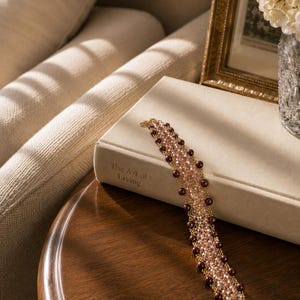 May include: A beaded bracelet featuring a woven lattice design of light pink crystals and dark brown round beads. The bracelet is constructed with gold-toned wire and is resting on a beige book titled The Art of Living, showcasing a detailed texture.