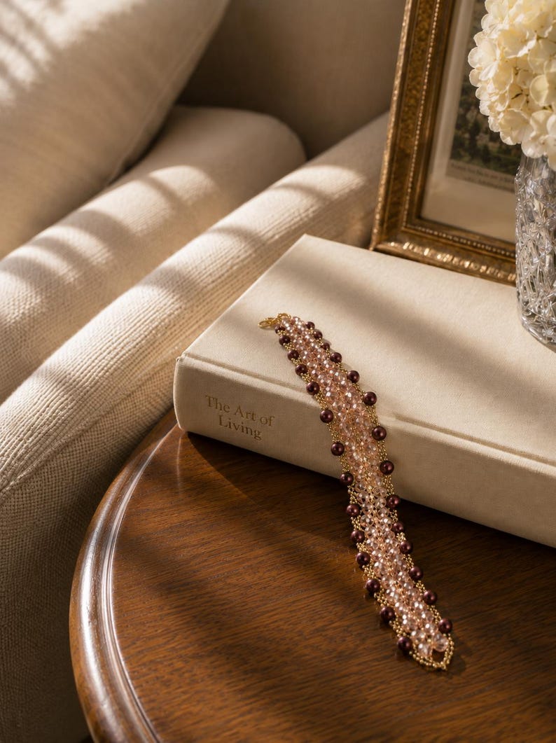 May include: A beaded bracelet featuring a woven lattice design of light pink crystals and dark brown round beads. The bracelet is constructed with gold-toned wire and is resting on a beige book titled The Art of Living, showcasing a detailed texture.