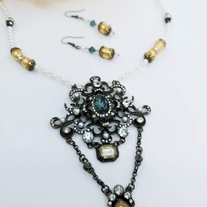 May include: An antique-style jewelry set featuring a necklace and matching earrings. The necklace has a large, ornate pendant with a central teal stone, surrounded by clear crystals and pearls. The earrings have a similar design with teal and yellow accents.
