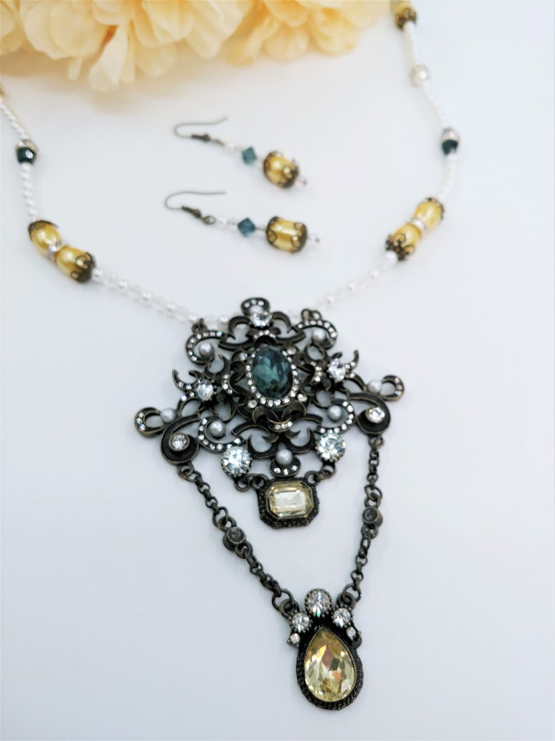 May include: An antique-style jewelry set featuring a necklace and matching earrings. The necklace has a large, ornate pendant with a central teal stone, surrounded by clear crystals and pearls. The earrings have a similar design with teal and yellow accents.