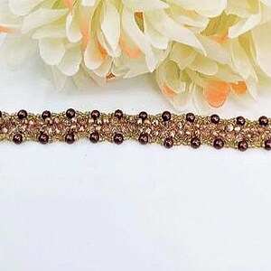 May include: A beaded bracelet featuring a woven design of small, dark maroon spherical beads and light pink faceted crystal beads. The bracelet is constructed with a gold-toned thread or wire, creating a textured, intricate pattern throughout the band.