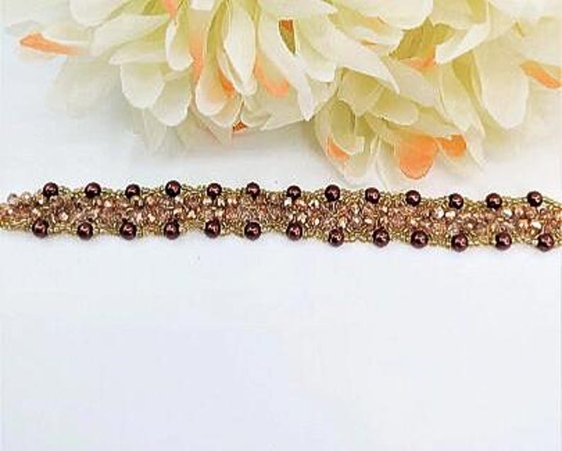 May include: A beaded bracelet featuring a woven design of small, dark maroon spherical beads and light pink faceted crystal beads. The bracelet is constructed with a gold-toned thread or wire, creating a textured, intricate pattern throughout the band.