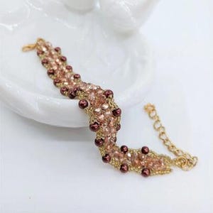 May include: A beaded bracelet featuring a woven pattern of light pink faceted crystals and dark maroon round beads. The bracelet is constructed with gold-toned thread and includes a gold-toned metal chain extender with a lobster claw clasp.
