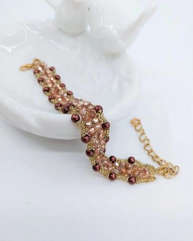 May include: A beaded bracelet featuring a woven pattern of light pink faceted crystals and dark maroon round beads. The bracelet is constructed with gold-toned thread and includes a gold-toned metal chain extender with a lobster claw clasp.