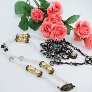 May include: An ornate necklace with a large, dark metal pendant adorned with clear crystals and a central blue stone. The necklace features strands of small white pearls and decorative gold-toned beads. Pink roses are in the background.