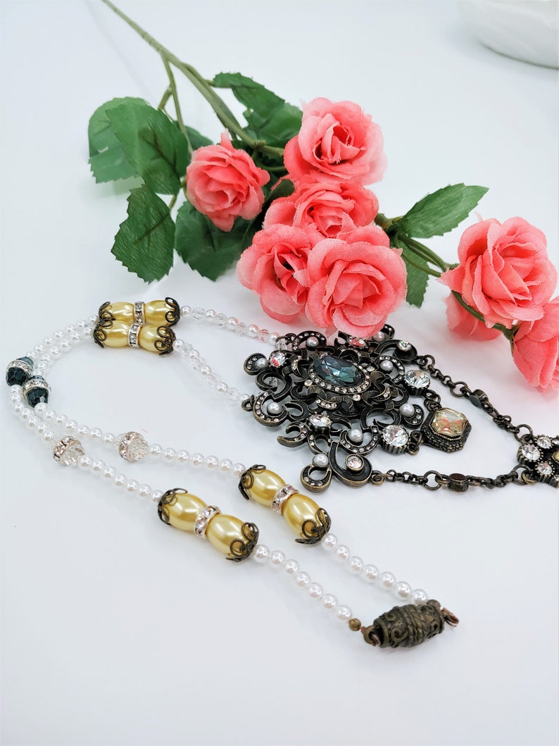 May include: An ornate necklace with a large, dark metal pendant adorned with clear crystals and a central blue stone. The necklace features strands of small white pearls and decorative gold-toned beads. Pink roses are in the background.