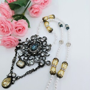 May include: An ornate, vintage-style necklace with a large, dark metal pendant featuring a teal-colored stone and clear crystals. The necklace has strands of faux pearls and cream-colored beads. Pink roses are in the background.