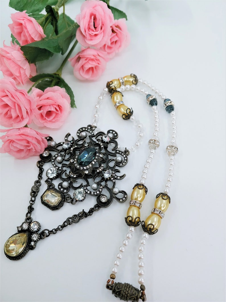 May include: An ornate, vintage-style necklace with a large, dark metal pendant featuring a teal-colored stone and clear crystals. The necklace has strands of faux pearls and cream-colored beads. Pink roses are in the background.