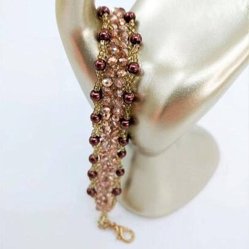 May include: A beaded bracelet featuring a central band of light pink faceted crystals flanked by two rows of dark maroon round beads. The beads are woven together with gold-toned thread, and the bracelet includes a gold-toned lobster claw clasp.