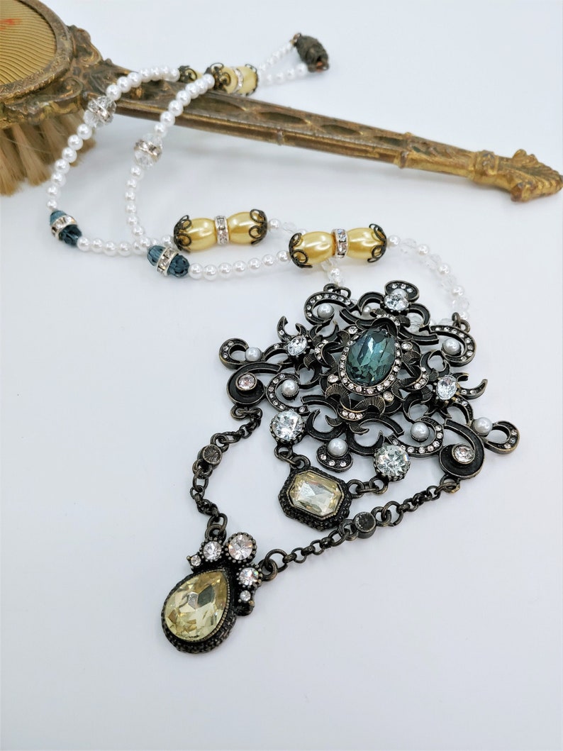 May include: An ornate, vintage-style necklace with a large central pendant featuring a teal-colored oval stone and clear crystals. The necklace has a dark metal finish, with smaller clear crystals and faux pearls. A vintage hand mirror is in the background.