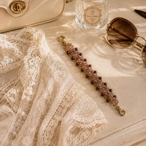 May include: A beaded bracelet featuring a gold-tone chain, small translucent light purple crystals, and dark purple round beads. The bracelet has a lobster claw clasp and is positioned on a surface next to a cream lace garment, a perfume bottle, and sunglasses.