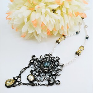 May include: An ornate, antique-style necklace with a large, dark metal pendant featuring a central teal-colored stone and smaller clear and pearl accents. The necklace has a delicate chain with pearl beads and yellow-gold accents. A bouquet of cream-colored flowers is in the background.