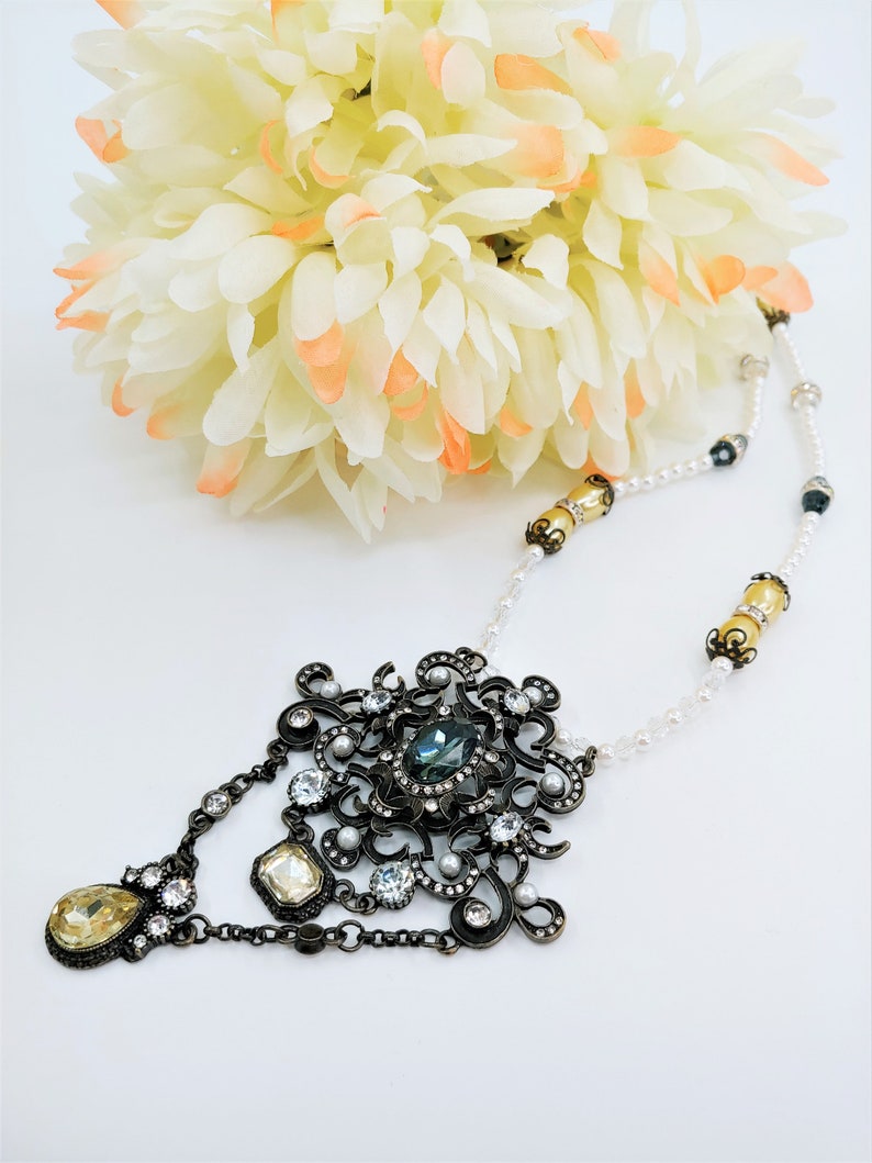 May include: An ornate, antique-style necklace with a large, dark metal pendant featuring a central teal-colored stone and smaller clear and pearl accents. The necklace has a delicate chain with pearl beads and yellow-gold accents. A bouquet of cream-colored flowers is in the background.