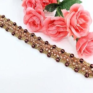 May include: A beaded bracelet featuring a woven pattern of small gold seed beads, light pink faceted crystals, and dark brown round beads. The bracelet is arranged in a diagonal line across the frame, accompanied by three artificial pink roses.