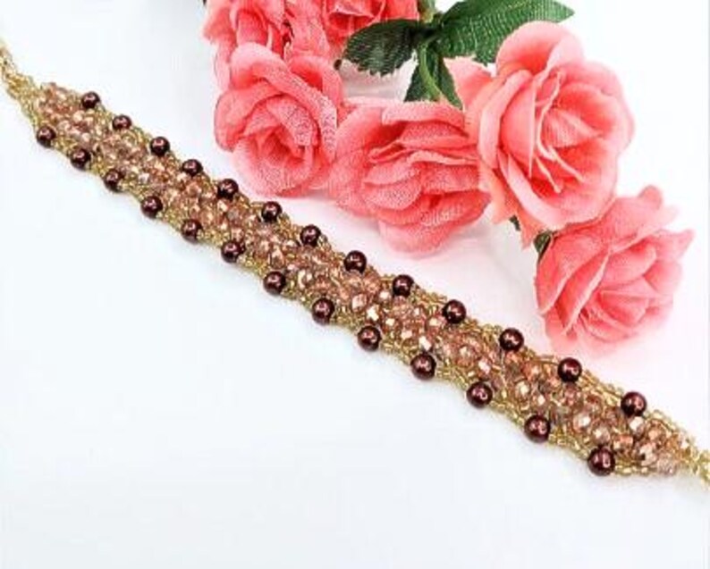 May include: A beaded bracelet featuring a woven pattern of small gold seed beads, light pink faceted crystals, and dark brown round beads. The bracelet is arranged in a diagonal line across the frame, accompanied by three artificial pink roses.