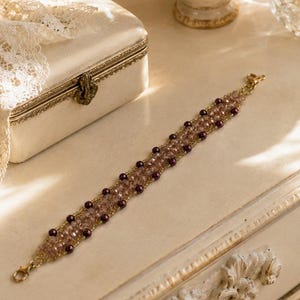 May include: A beaded bracelet featuring a central band of light pink faceted crystals flanked by rows of small, dark purple round beads. The design is held together by thin gold-toned wire and includes a gold-toned lobster claw clasp at each end.