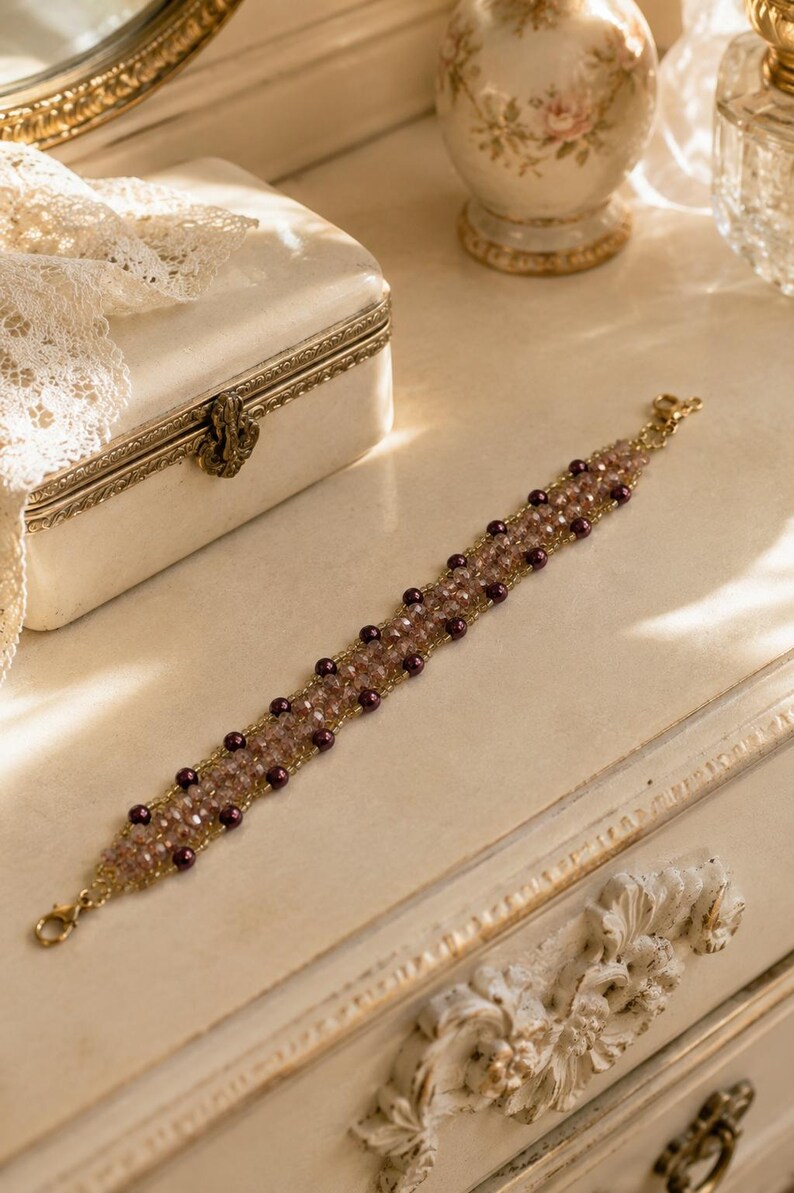 May include: A beaded bracelet featuring a central band of light pink faceted crystals flanked by rows of small, dark purple round beads. The design is held together by thin gold-toned wire and includes a gold-toned lobster claw clasp at each end.