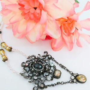 May include: An ornate necklace with a large, dark-colored central stone and smaller clear and yellow stones. The necklace has a detailed, antique-style design with a pearl strand and is set against a backdrop of pink flowers.
