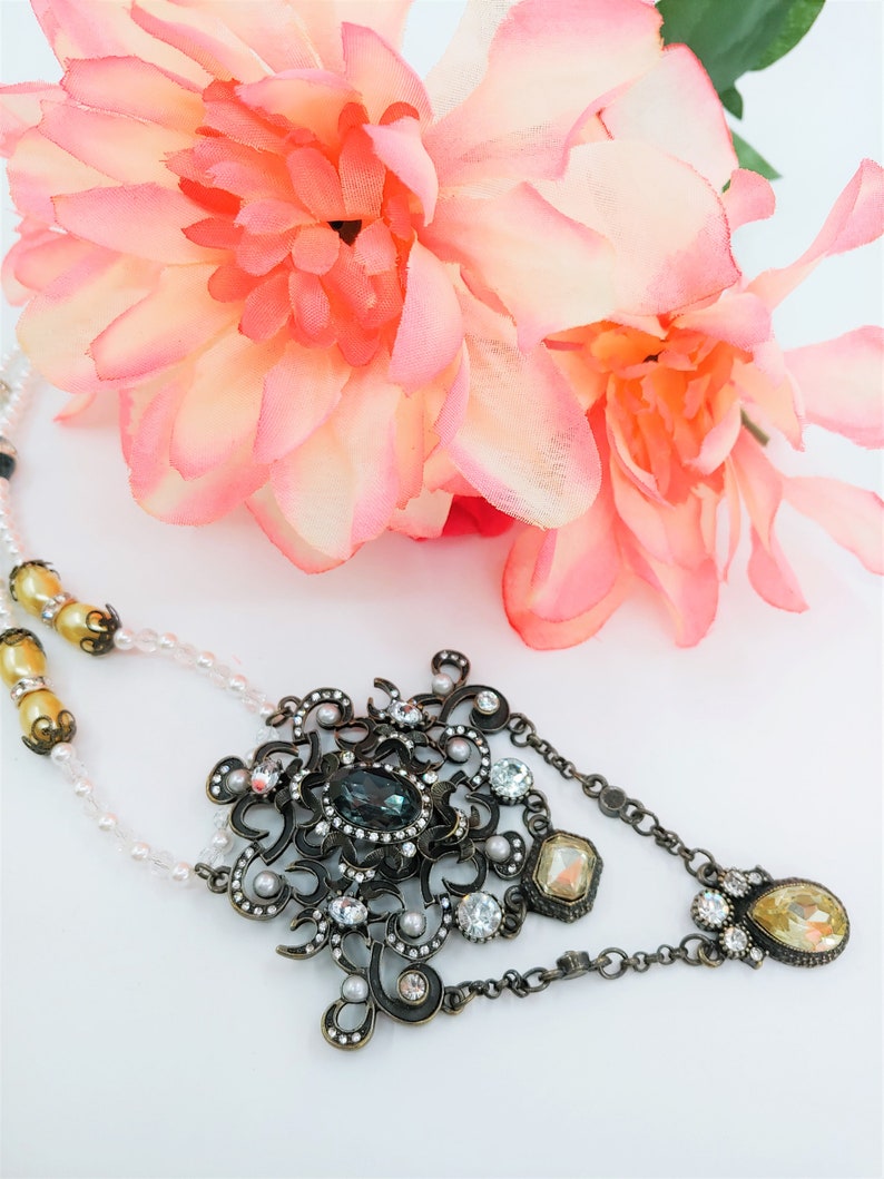 May include: An ornate necklace with a large, dark-colored central stone and smaller clear and yellow stones. The necklace has a detailed, antique-style design with a pearl strand and is set against a backdrop of pink flowers.