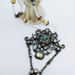May include: An ornate necklace featuring a large pendant with a teal-colored stone and smaller clear and yellow stones. The necklace has a pearl and bead strand with decorative accents. The metal has an antique bronze finish.