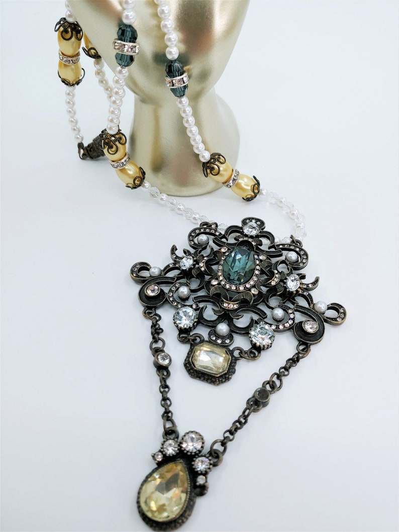 May include: An ornate necklace featuring a large pendant with a teal-colored stone and smaller clear and yellow stones. The necklace has a pearl and bead strand with decorative accents. The metal has an antique bronze finish.