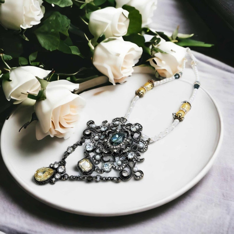 May include: An ornate, vintage-style necklace with a large central pendant featuring a teal-colored stone, surrounded by smaller clear and pearl-like accents. The necklace has a dark metal finish and is displayed on a white plate with white roses in the background.