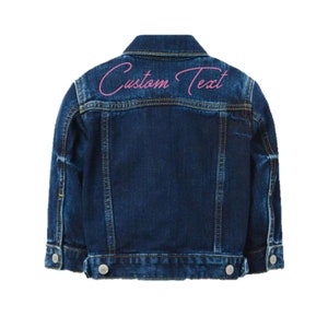 May include: A blue denim jacket with a pink embroidered text that says "Custom Text" on the back.