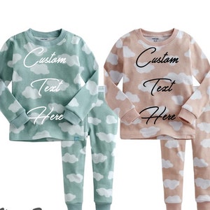 May include: Two sets of pajamas with a cloud print. One set is teal with white clouds and the other is pink with white clouds. Both sets have the text "Custom Text Here" on the shirts.