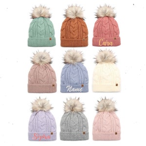 May include: Nine knitted winter hats with faux fur pom poms in various colors, including green, brown, pink, blue, white, lavender, and gray. Some hats have names embroidered on them, such as "Cara", "Name", and "Sophia".